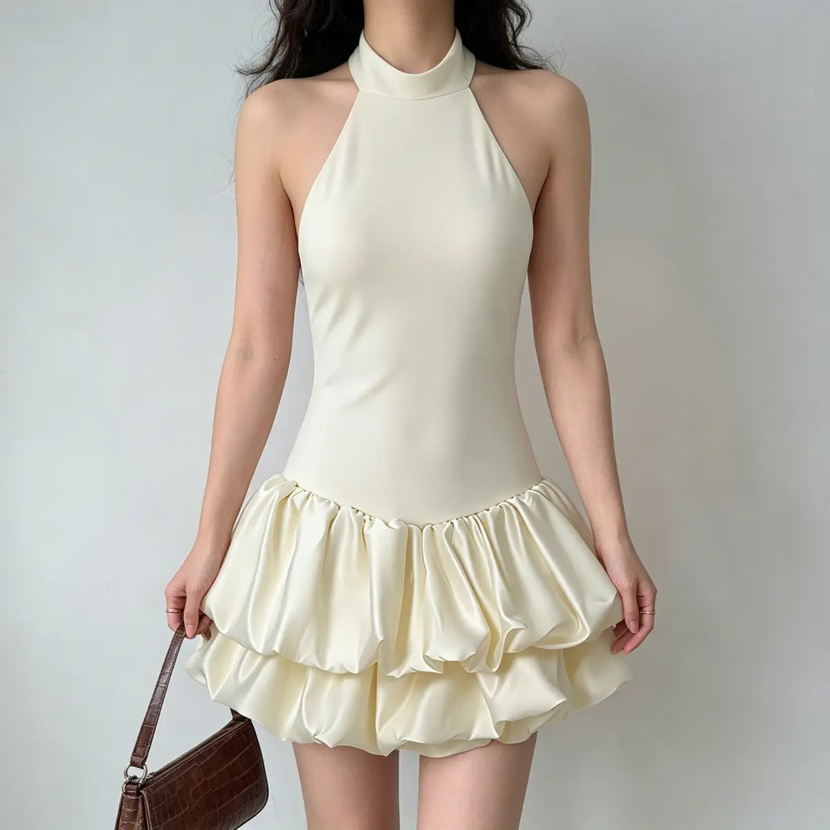 Full front view of a model wearing a cream halter-neck puffball mini dress with double-layered skirt, holding a brown crocodile bag.