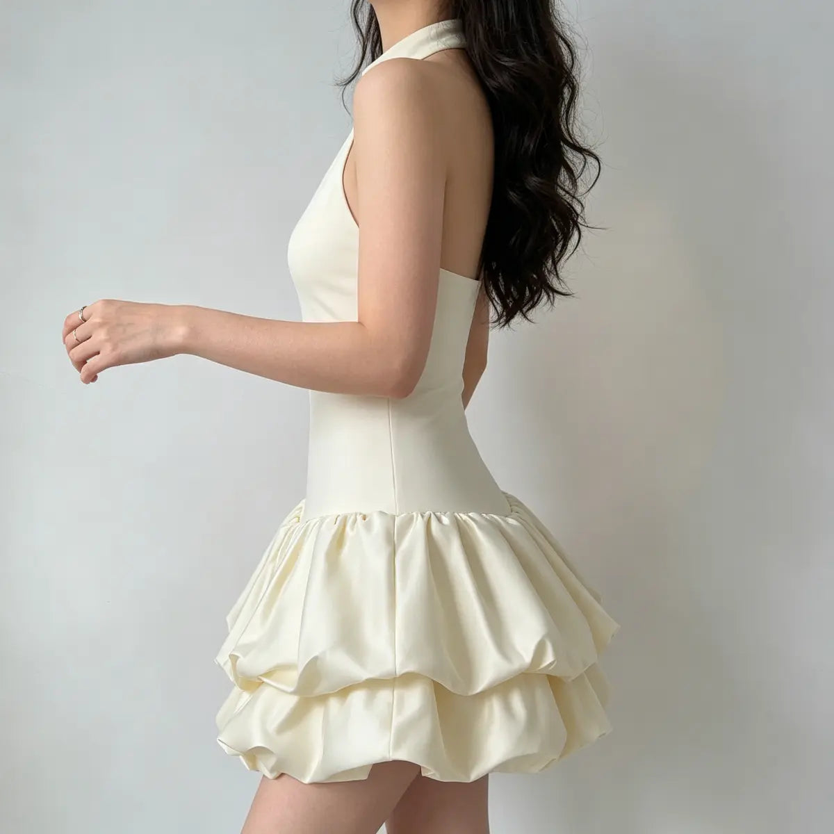 Profile view of a model wearing a cream halter-neck puffball mini dress, highlighting the fitted bodice and puffy skirt.