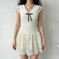 Model wearing a cream lace mini dress with a large flat collar, cap sleeves, and a black ribbon bow, holding a small wicker bag and wearing a pearl necklace.