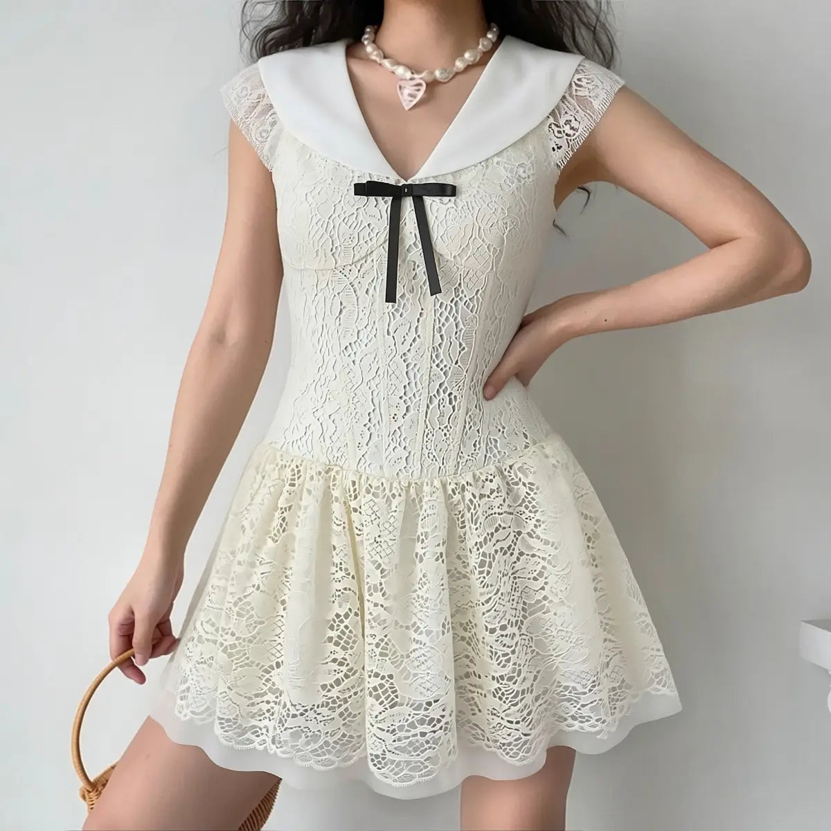 Front view of a model wearing a cream lace mini dress with a large flat collar, cap sleeves, and a black ribbon bow, wearing a pearl necklace.