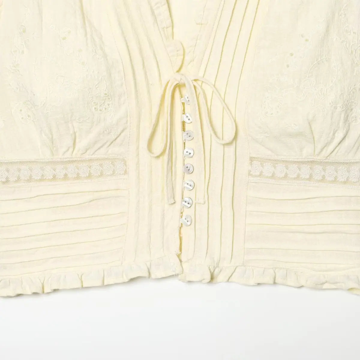 Front view of the cream bustier top, showing the V-neckline, tie detail, shell buttons, and lace/embroidery accents