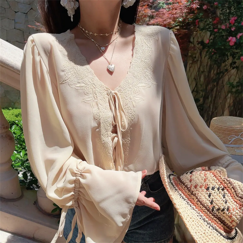 Outdoor shot of a model wearing a cream lace-trim chiffon blouse with front tie detail, holding a woven hat.