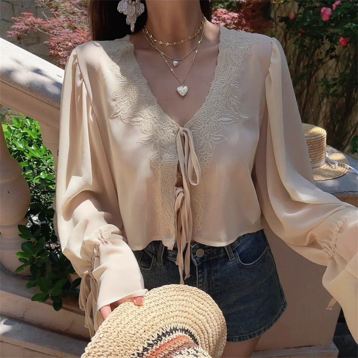 Styled shot of a model wearing a cream lace-trim chiffon blouse with denim shorts, holding a woven hat.