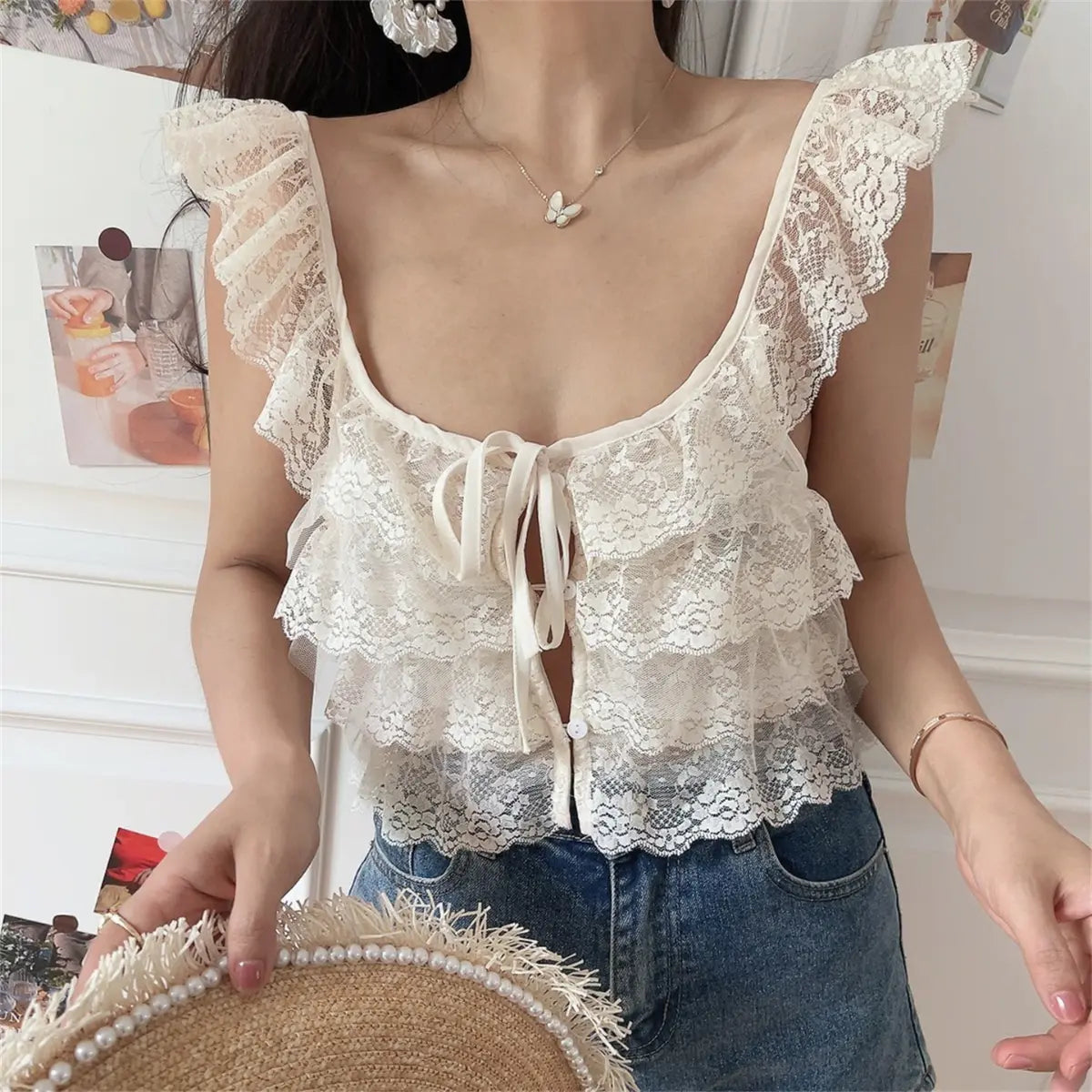 Front view of a woman wearing a cream layered lace camisole with a front tie and buttons, paired with jeans and holding a straw hat.