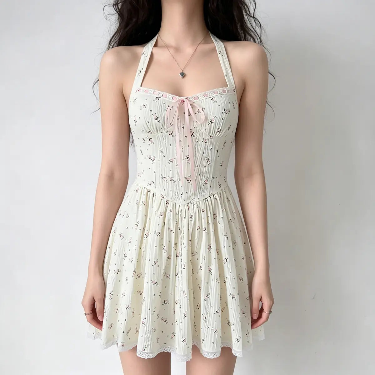  Model wearing a cream halter-neck mini dress with a pink floral print, a pink ribbon tie at the bust, and lace trim at the hem.