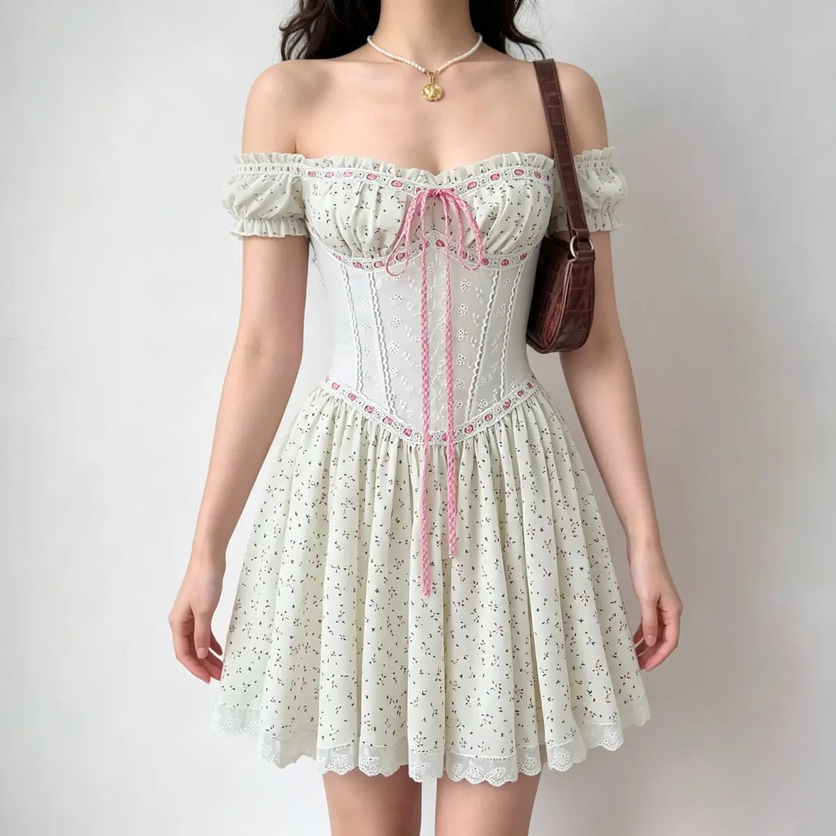Model wearing an off-the-shoulder cream dress with a pink floral print, lace-up front, and puffed sleeves, carrying a brown handbag.
