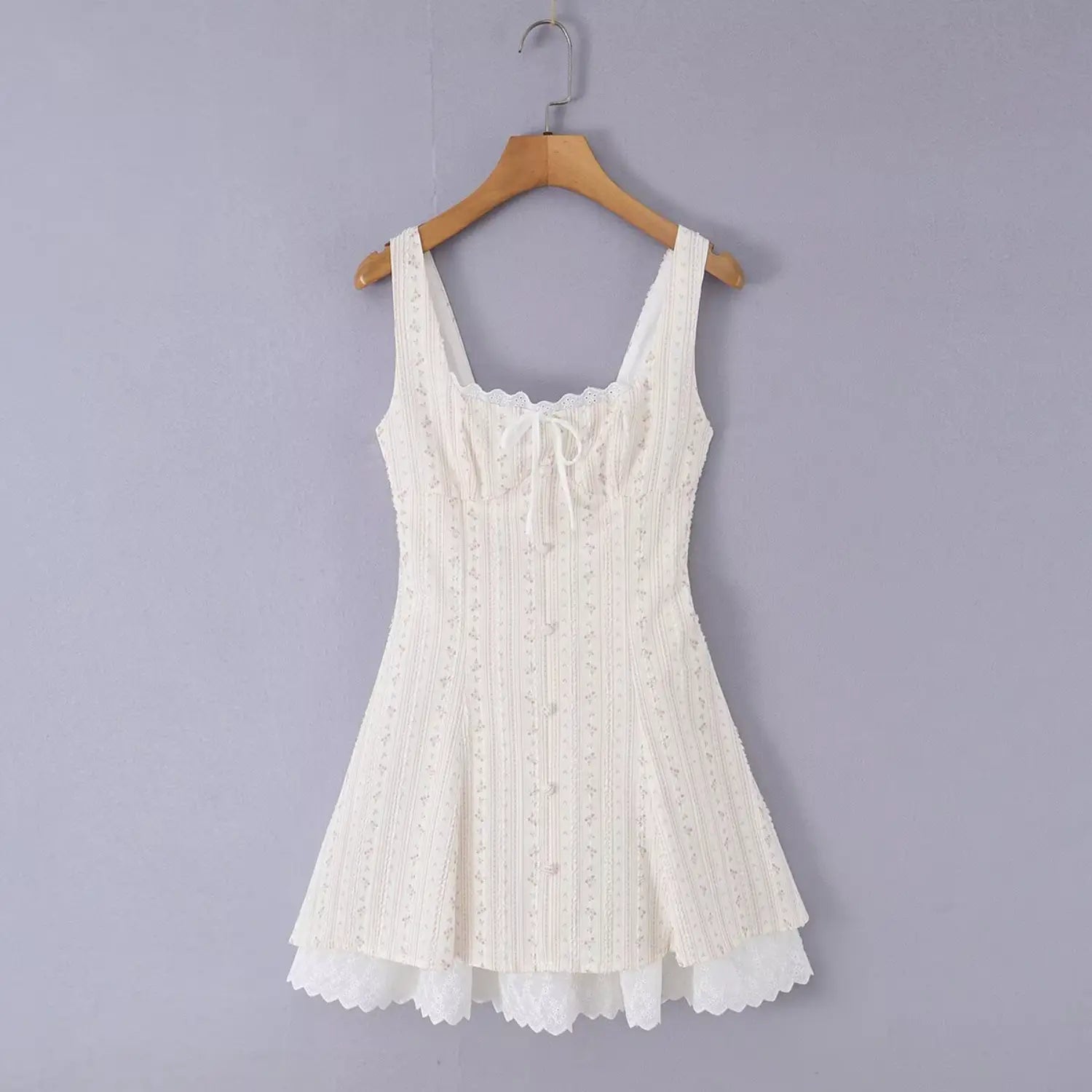 Hanger shot of cream mini cami dress with tiny pink vertical floral print, square lace trim neck, front fabric tie bow, faux bust buttons, layered lace under slip hem, cottagecore slim fit sundress