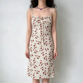 Model wearing a cream midi camisole dress with a red floral print, a gathered bust, and a front tie detail.