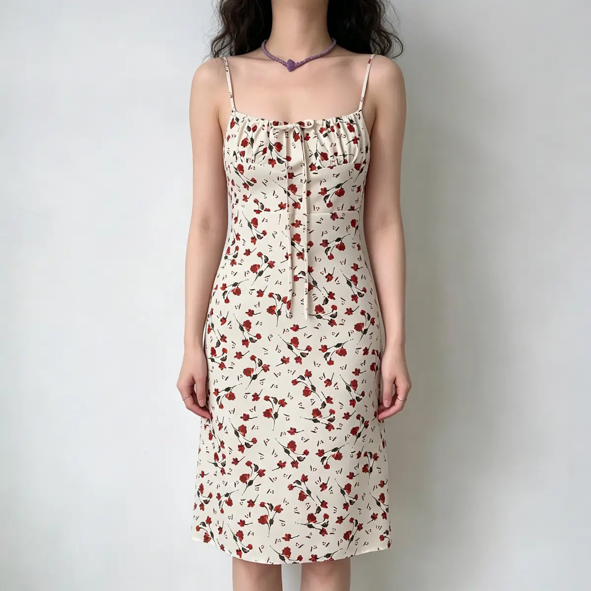 Model wearing a cream midi camisole dress with a red floral print, a gathered bust, and a front tie detail.