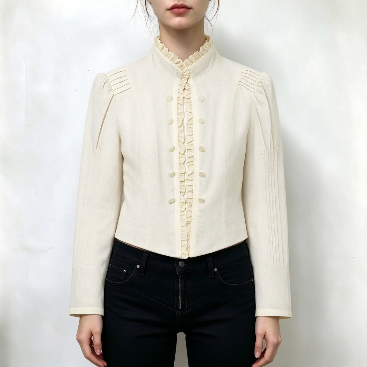 Model wearing cream cropped blouse, ruffled mandarin collar, double row buttons, front ruffle placket, pleated puffed long sleeves, tailored slim fit, paired with black jeans, plain white background