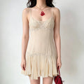 Model wearing a cream-colored sheer slip mini dress with intricate lace trim at the bust, waist, and hem, styled with a red handbag and red pendant necklace.