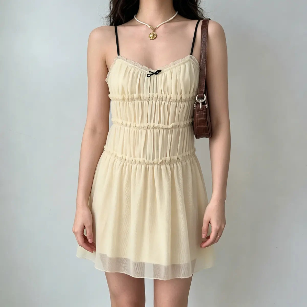  Full front view of a model wearing a cream smocked chiffon mini dress with ruffled tiers and a small black bow.