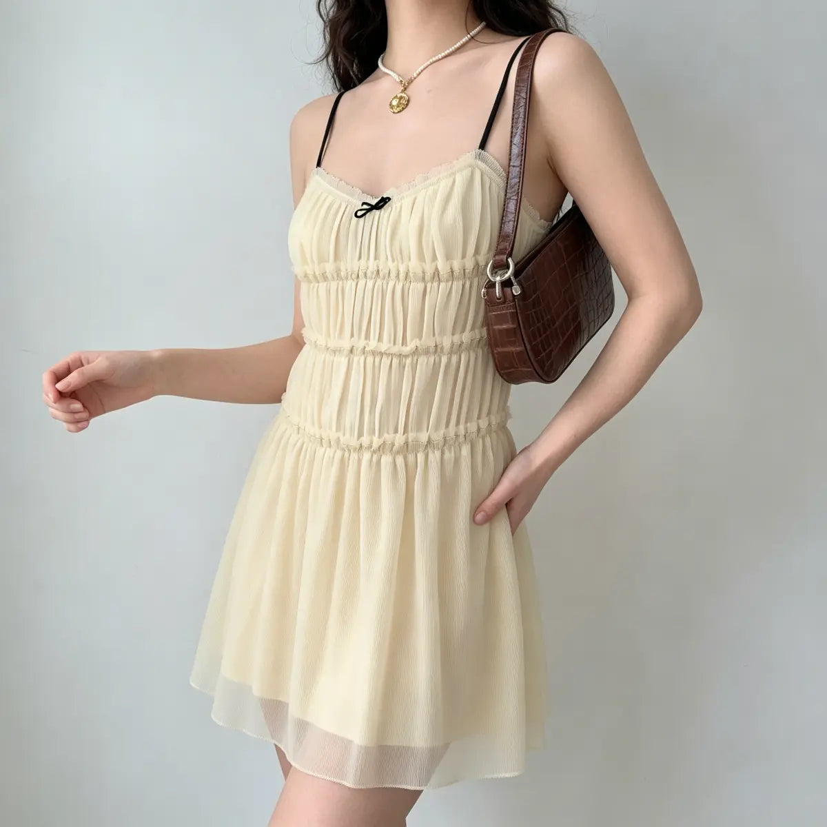 Model posing with one hand on her hip, wearing a cream smocked chiffon mini dress with black straps.