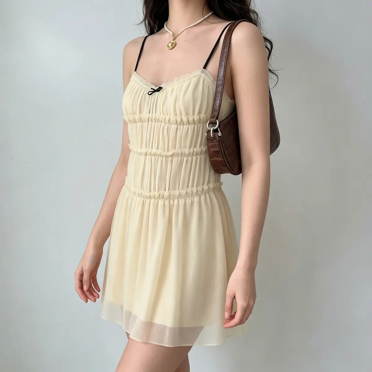 Side view of a model wearing a cream smocked chiffon mini dress with black spaghetti straps, carrying a brown crocodile bag.