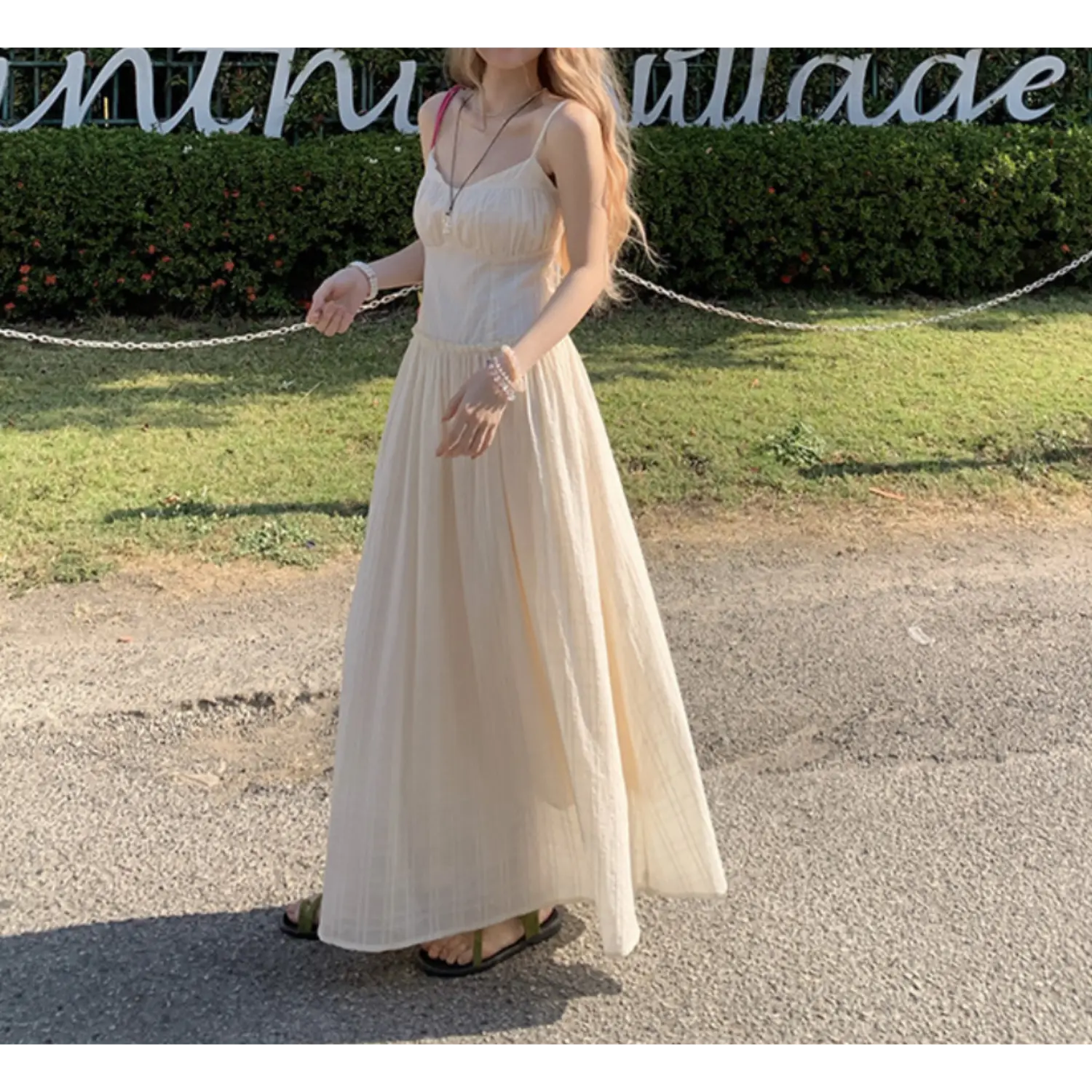 Three-quarter outdoor lawn lifestyle shot of cream textured cami maxi dress, fine vertical waffle fabric, thin adjustable spaghetti straps, ruched sweetheart bust, ruffled empire waist seam, full flowy A-line floor skirt, styled with green strap slide sandals, green hedge & vintage letter signage backdrop