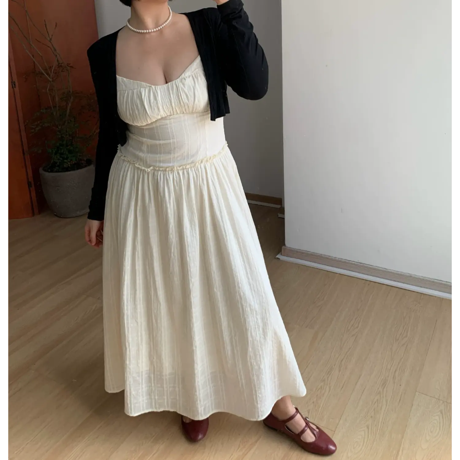 Indoor mirror layered styling shot of waffle knit cami maxi dress matched with slim long sleeve black cropped knit cardigan, sweetheart ruched bust, delicate ruffle waist trim, full flowy floor skirt, burgundy mary jane shoes, minimalist light wood floor backdrop