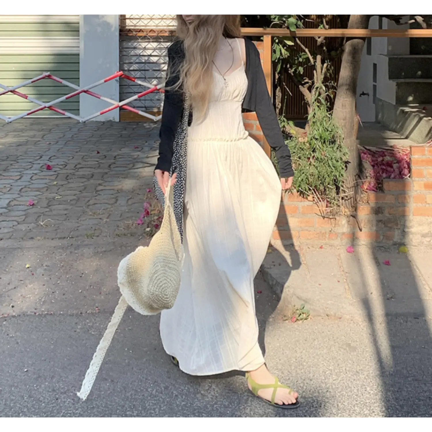 Relaxed outdoor street walking shot of fitted empire waist cami maxi sundress, subtle vertical waffle texture fabric, thin adjustable cami straps, hidden side pocket (model resting one hand inside pocket), straw woven shoulder bag, green slide sandals, residential brick & foliage scenery