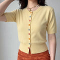 Lifestyle model wearing solid cream yellow fine knit cardigan, rib crew round neck, full vertical multi-color 3D daisy flower button placket, short rib cuff sleeves, slim cropped rib hem, matching orange textured bottom, shell starfish necklace outfit display