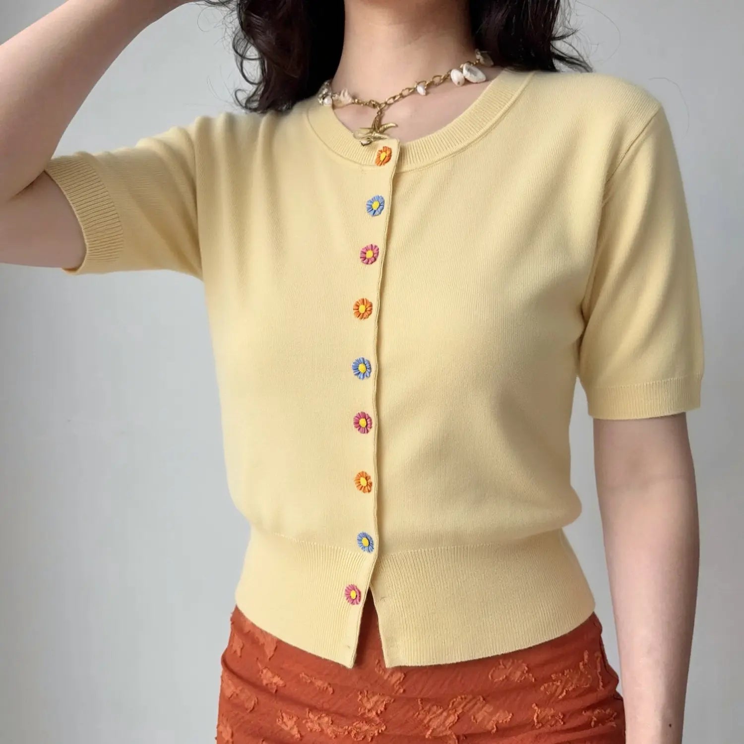 Lifestyle model wearing solid cream yellow fine knit cardigan, rib crew round neck, full vertical multi-color 3D daisy flower button placket, short rib cuff sleeves, slim cropped rib hem, matching orange textured bottom, shell starfish necklace outfit display