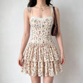 Model wearing a cream corset-style mini dress with a yellow and red floral print, tiered ruffled skirt, and thin tie straps, carrying a brown handbag.