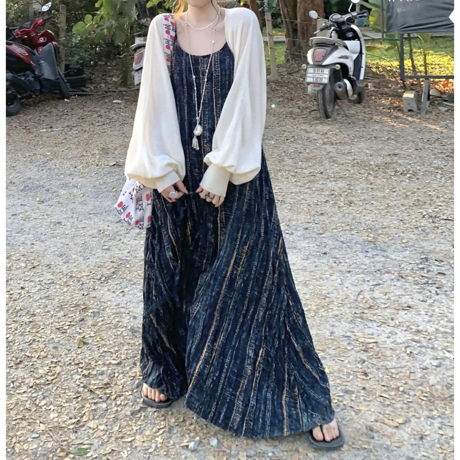 Outdoor full body layered styling shot of textured striped slip maxi dress, paired with loose white waffle knit balloon sleeve cropped cardigan, thin matching choker tie, canvas print shoulder bag, black slide sandals, rural roadside backdrop with motorbikes & dry foliage