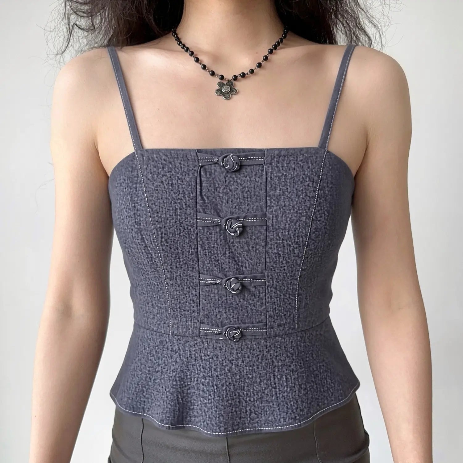 Lifestyle model wearing dark denim frog button camisole, thin shoulder straps, fitted peplum cropped hem, matched dark gray trousers & vintage flower pendant necklace outfit display