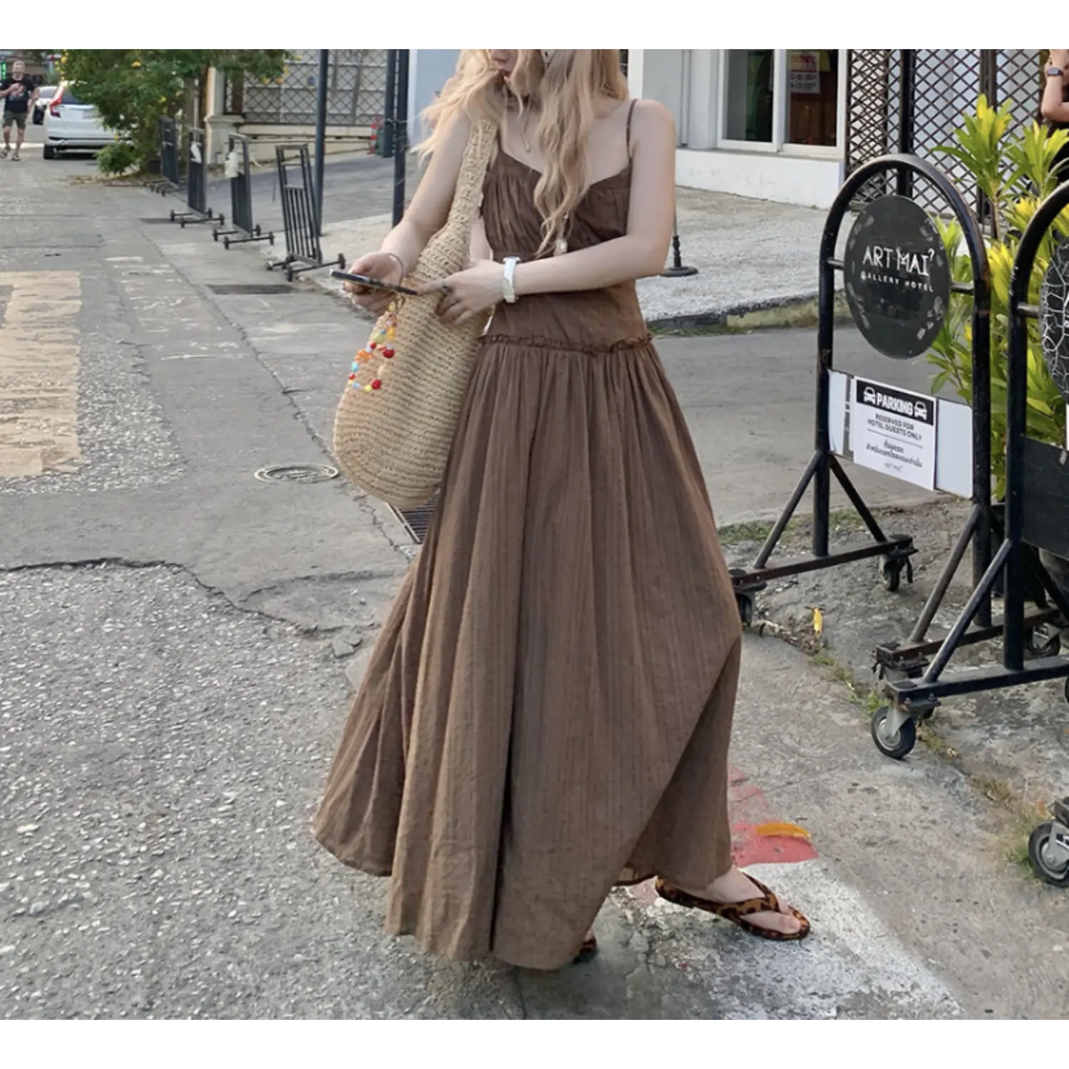 Three-quarter roadside walking lifestyle shot of coffee brown textured slip maxi dress, fitted ruched bust, ruffle high waist separating slim bodice & sweeping flowy skirt, thin cami straps, oversized straw tote bag, leopard slide sandals, vintage hotel metal signage & brick pavement background