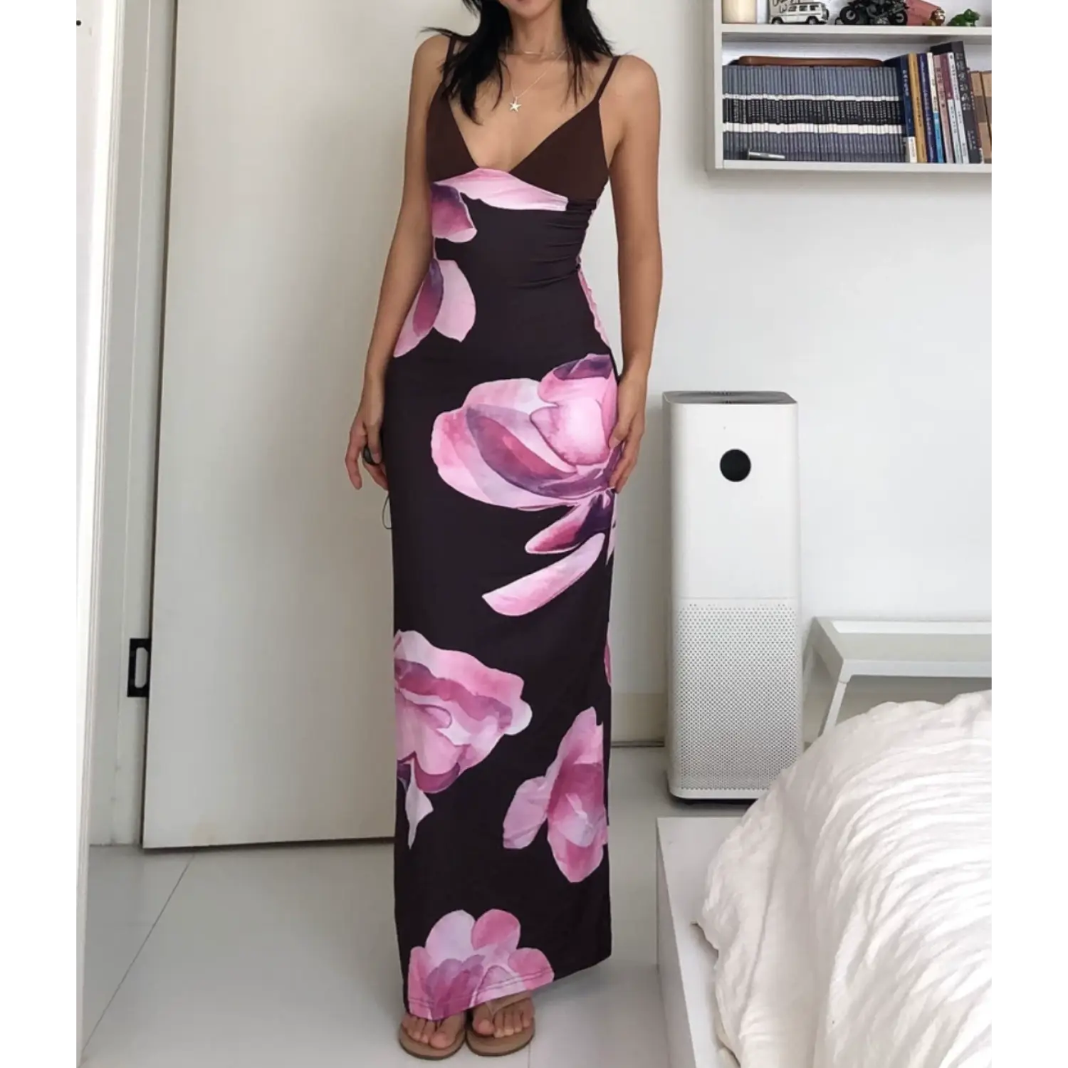 Medium full indoor bedroom lifestyle shot of V-neck stretch slip maxi dress, model resting one hand on hip to highlight ruched waist fit, soft gradient pink magnolia floral print all over dark brown fabric