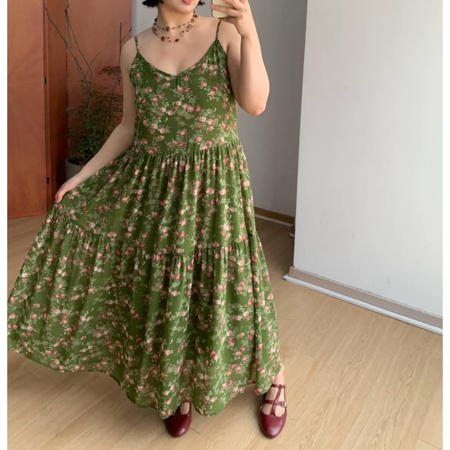 Angled indoor mirror lifestyle photo of forest green pink rose tier slip maxi dress, model lifting skirt hem to show lightweight sheer chiffon fabric, burgundy two-strap mary janes