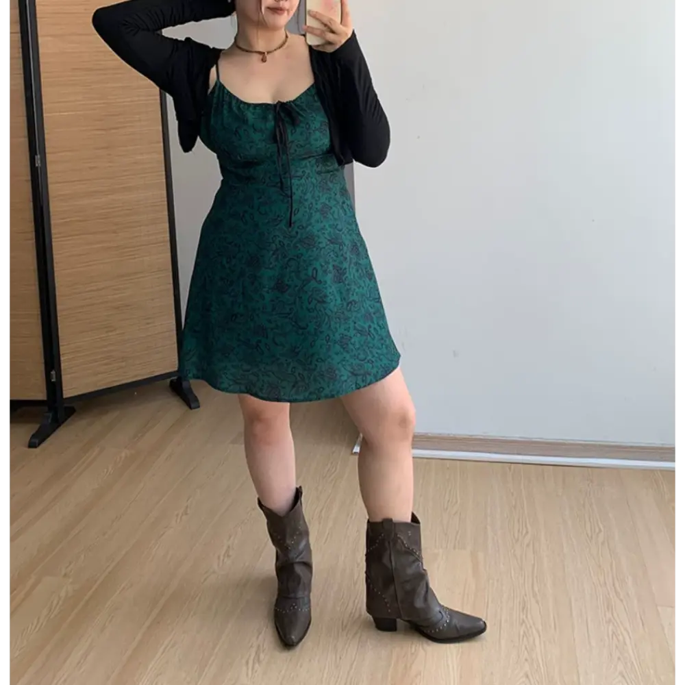 Full body indoor mirror shot of dark green paisley print spaghetti strap mini dress, layered slim black long sleeve cropped cardigan, paired with brown studded western ankle boots
