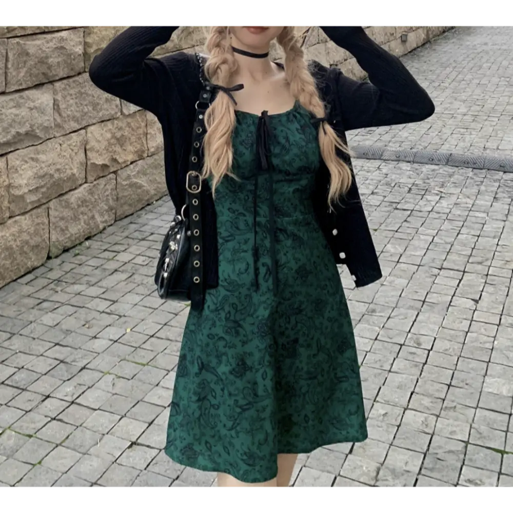 Full body outdoor street lifestyle shot of green paisley cami mini dress, layered black ribbed cropped cardigan, double blonde braids, carrying black metal eyelet crossbody bag