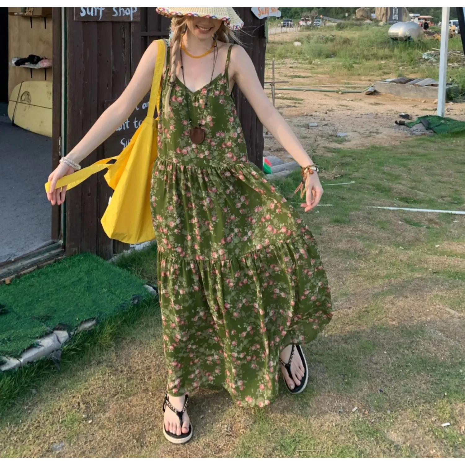 Seaside surf shop lawn lifestyle full shot of forest green ditsy rose tier slip maxi dress, model holding yellow tote bag strap, black chain platform flip flops, wooden shop backdrop