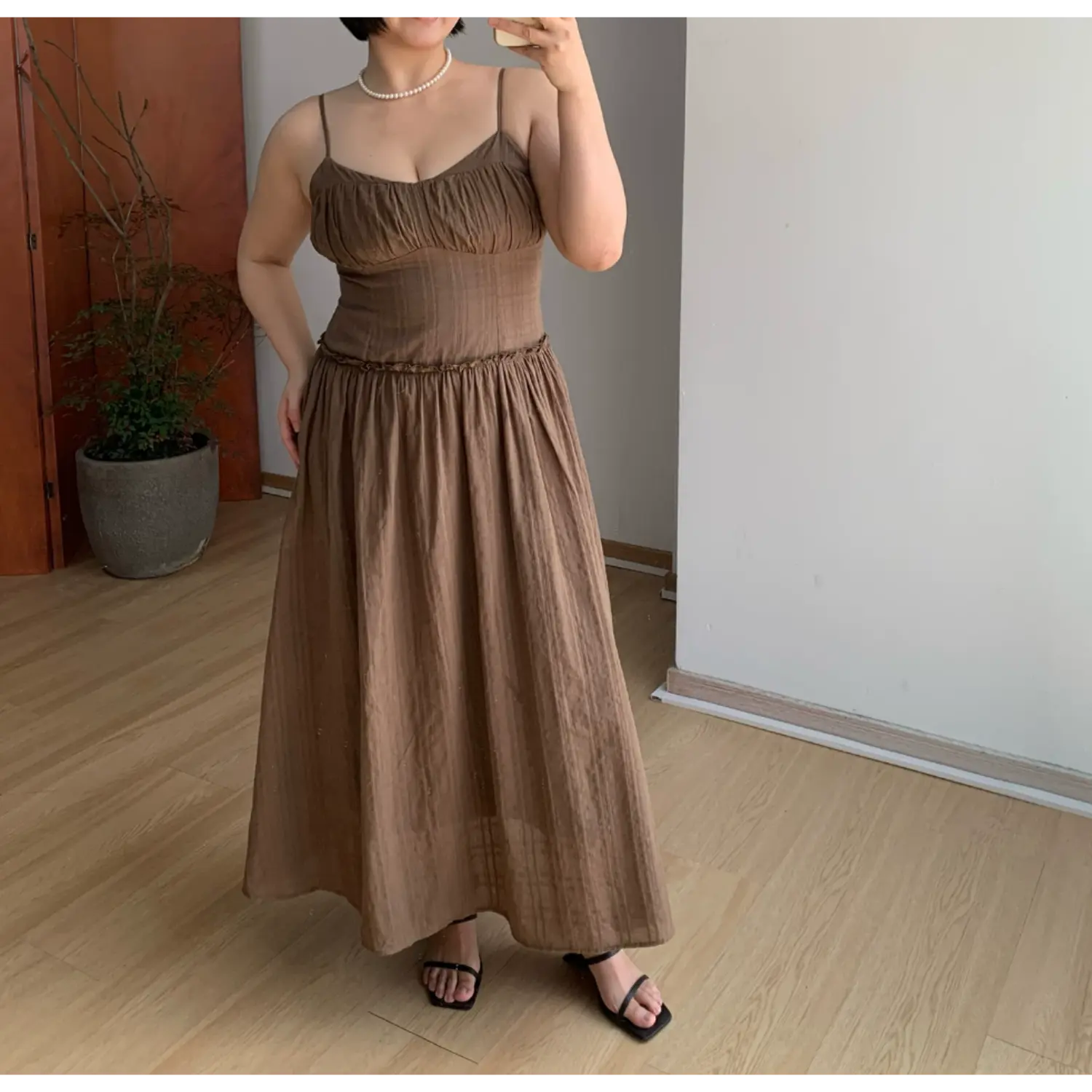 Full spread front indoor mirror shot of feminine fitted cami maxi sundress, ruched sweetheart bust, delicate ruffle high waist, extra wide flowy A-line floor skirt, subtle vertical textured fabric, thin spaghetti straps, black square slide sandals, plain white wall & light wood flooring decor