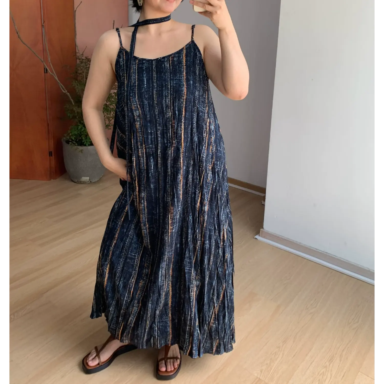 Indoor mirror full-body shot of loose crinkle maxi slip dress, dark navy base with faded vertical bronze & pale blue striped print, thin adjustable spaghetti straps, scoop neck, matching long fabric choker tie, flowy floor-length A-line skirt, styled with brown thong platform slide sandals on light wood floor