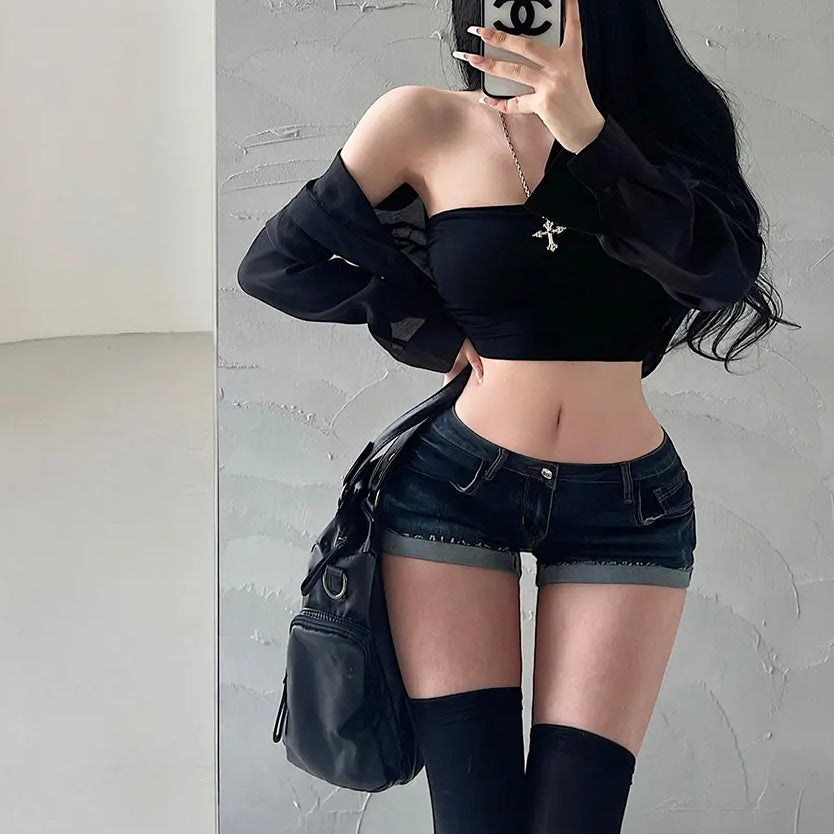 A woman wearing dark wash low-rise denim shorts, styled with a black off-shoulder top, sheer shirt, cat ear headband, and black thigh-high socks.