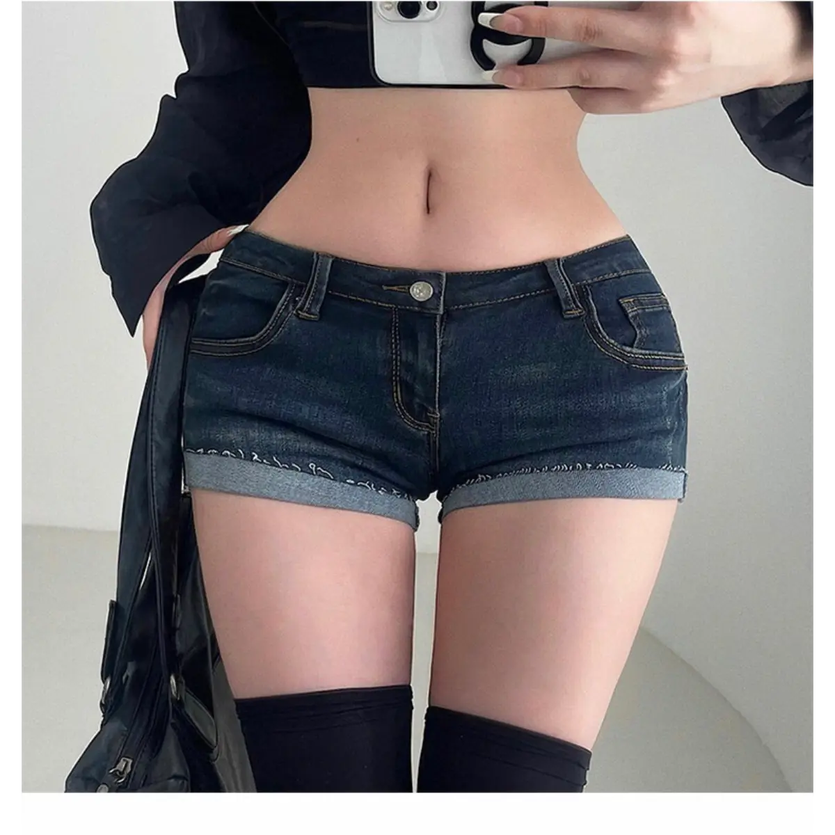 A woman wearing dark wash low-rise denim shorts with rolled cuffs, styled with a black crop top, sheer shirt, and black thigh-high socks.
