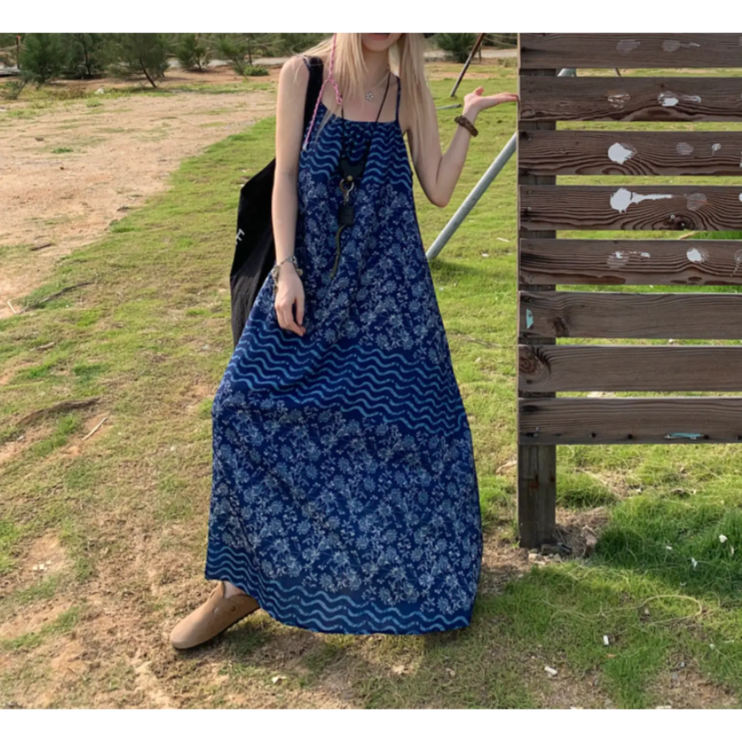 Static outdoor grass full shot of sleeveless flowy batik slip maxi dress, indigo floral + light blue wave panel print, thin spaghetti straps, loose unstructured A-line skirt, model resting one hand on wooden fence post, beige slip-on clogs, tropical dry grass scenery
