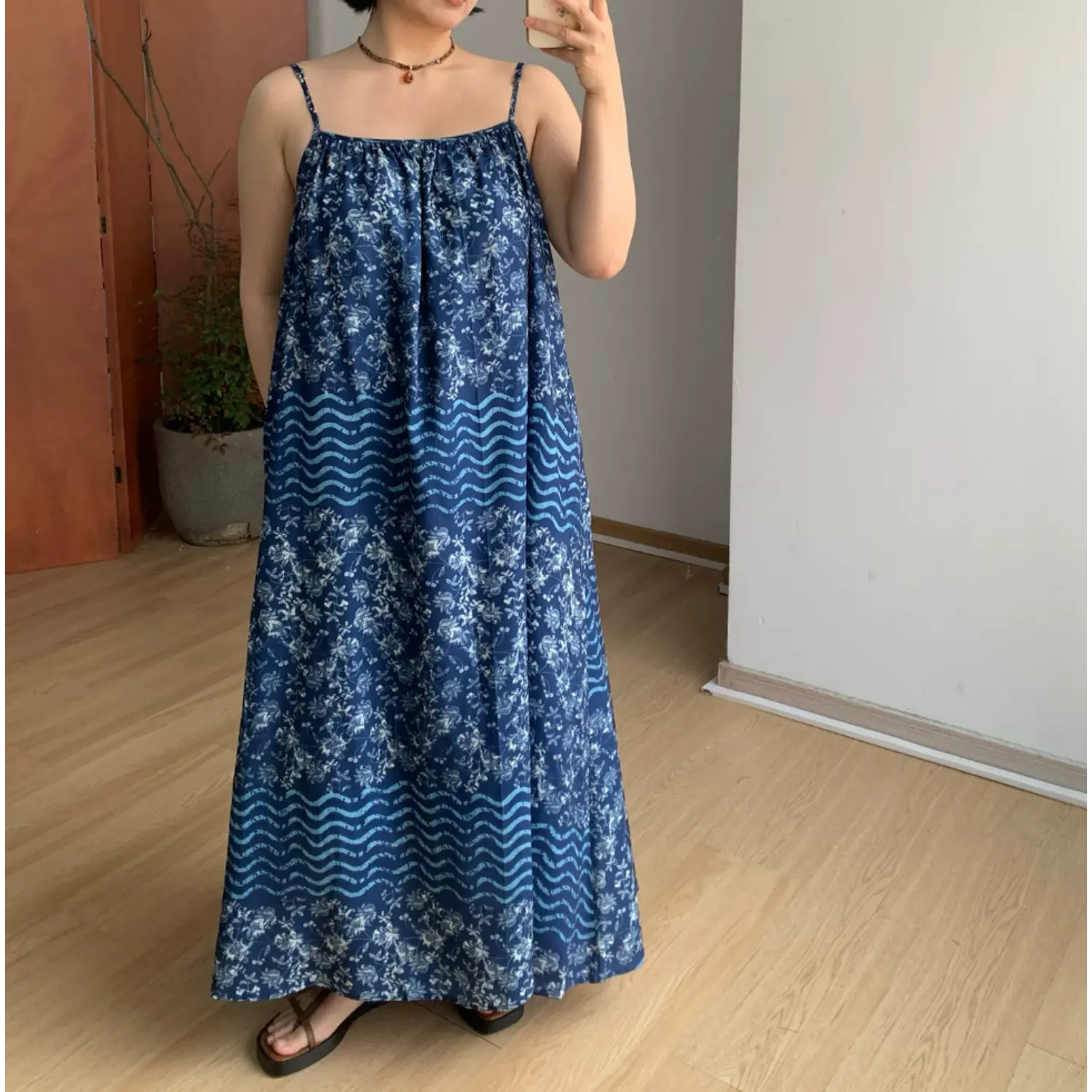 Indoor mirror full-body shot of oversized flowy cami maxi dress, deep navy blue base with batik white floral & light blue horizontal wave stripe panel print, thin adjustable spaghetti straps, gathered square neck bust, loose A-line swing floor skirt, hidden side pockets, styled with brown thong platform slide sandals on light wood flooring