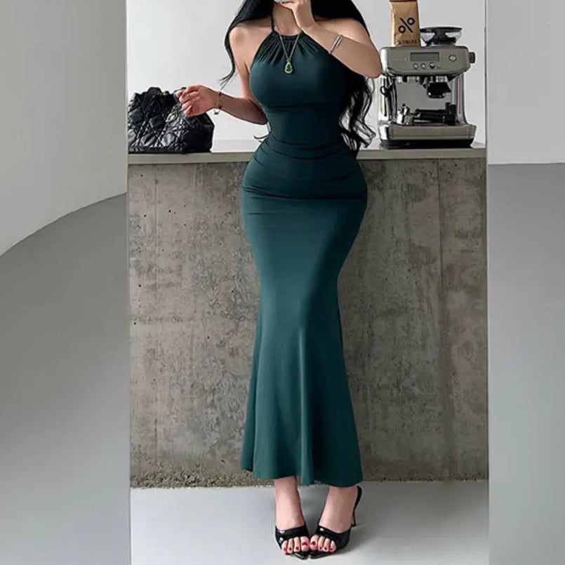 A woman wearing a deep green halter-neck mermaid maxi dress with ruched waist and bodycon fit, styled with black accessories.