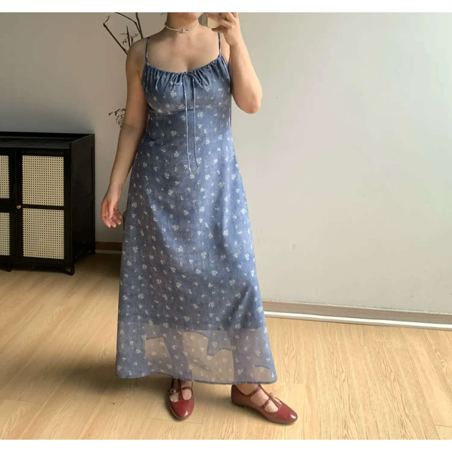 Indoor mirror full shot of faded denim blue tiny white floral slip maxi dress, thin adjustable straps, front bust self-fabric drawstring tie, paired with burgundy mary jane flats