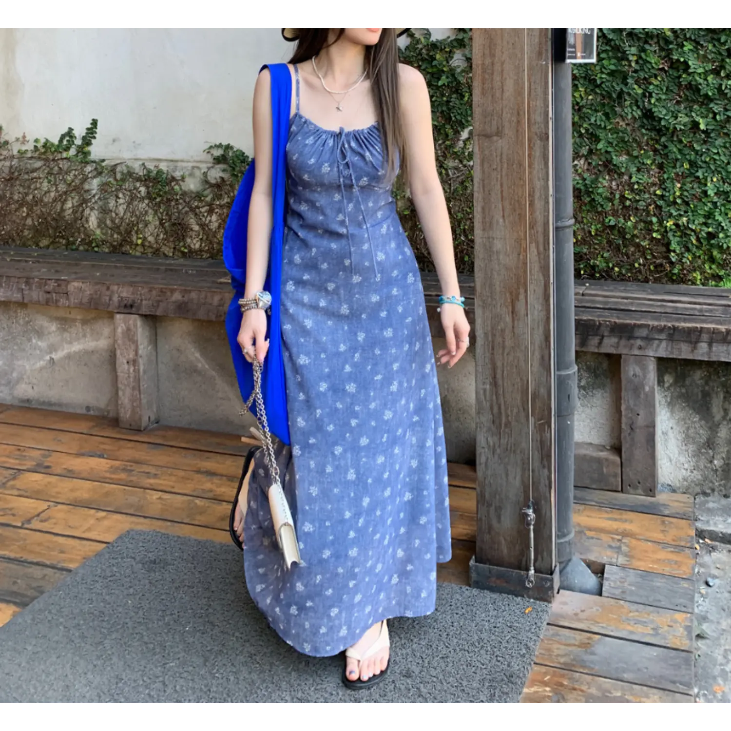 Outdoor wooden terrace three-quarter shot of faded blue tiny white floral maxi slip dress, model carrying oversized bright blue canvas tote and mini white leather chain bag, wearing simple slide sandals