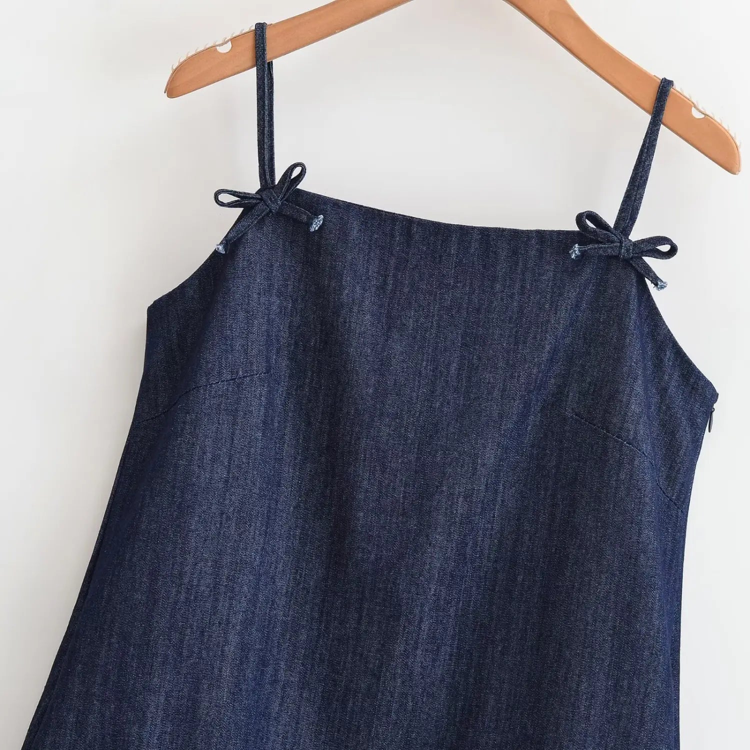 Hanging shot close‑up front bodice of dark denim cami dress, fabric bow tie details on spaghetti shoulder straps, side invisible zipper