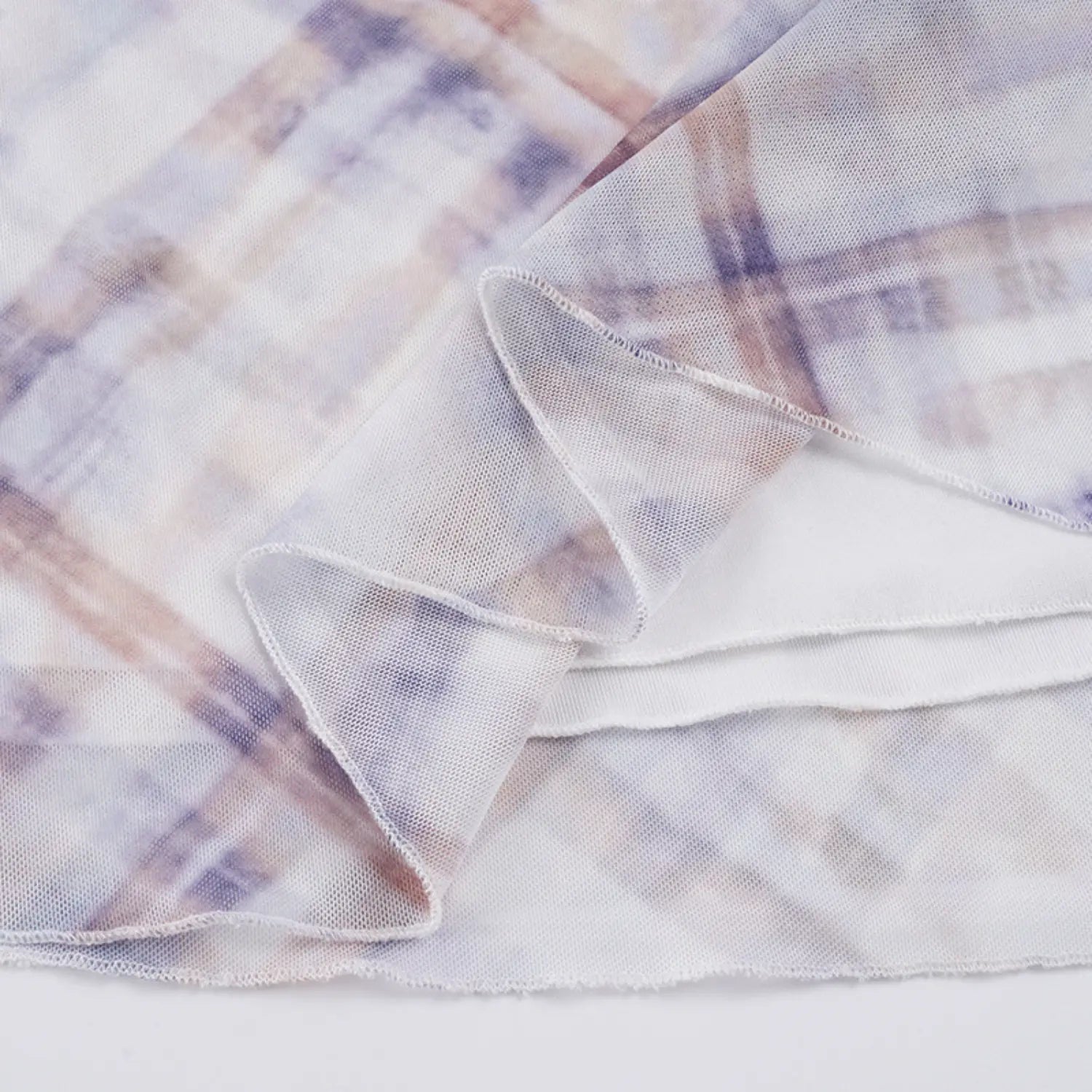 Close layered hem texture shot of two-tier mesh slip skirt, sheer printed outer mesh over solid white lining, clean wavy overlock stitching along raw fabric edges