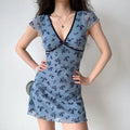 Model hands‑on‑hips pose dusty blue floral mesh bodycon mini dress v‑neck velvet bow cap sleeve ruffle lettuce hem