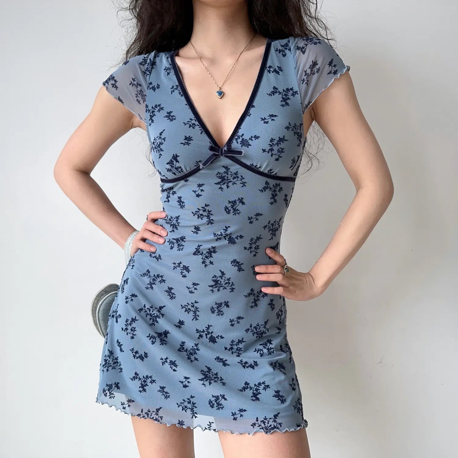 Model hands‑on‑hips pose dusty blue floral mesh bodycon mini dress v‑neck velvet bow cap sleeve ruffle lettuce hem