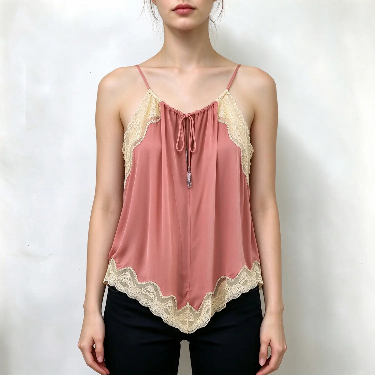 Model wearing dusty pink cami top with cream scalloped lace trim, tie front tassel detail, thin spaghetti straps, flowy asymmetrical hem, paired with black pants on plain white wall