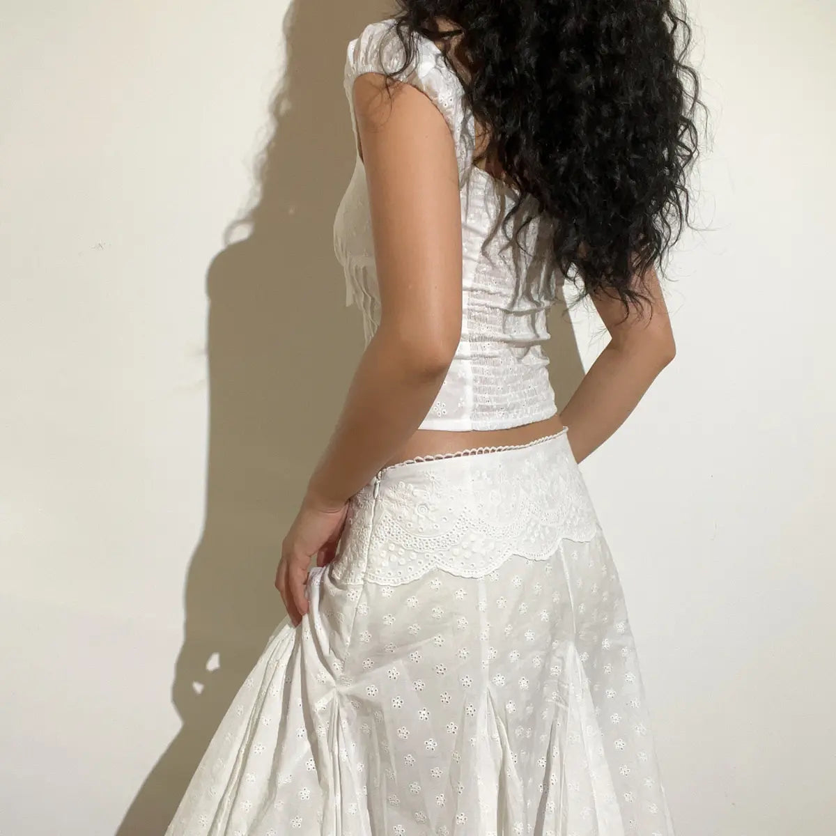 Back view of a white eyelet embroidered corset-style top and matching skirt, showing the smocked back panel of the top and lace-trimmed skirt hem.