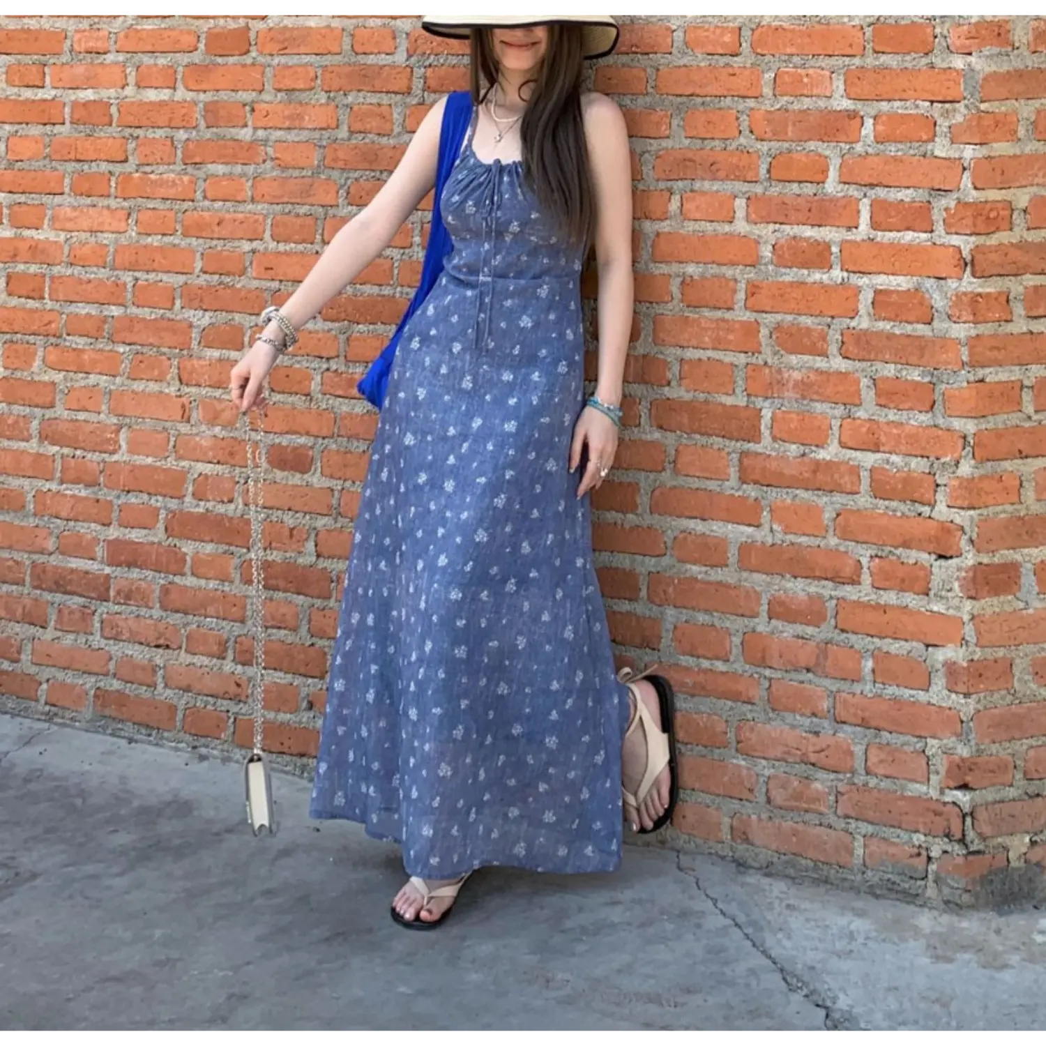 Urban street outdoor full shot of denim wash blue micro floral slip maxi dress, model leaning against red brick wall, straw wide brim hat, oversized blue tote bag and flat slide sandals