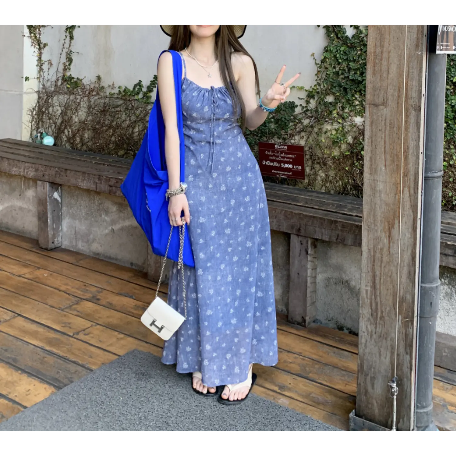 Vacation outdoor lifestyle shot of denim wash blue micro floral slip maxi dress, model making peace sign, accessorized with straw sun hat, bright blue tote bag and white chain crossbody bag, slide sandals