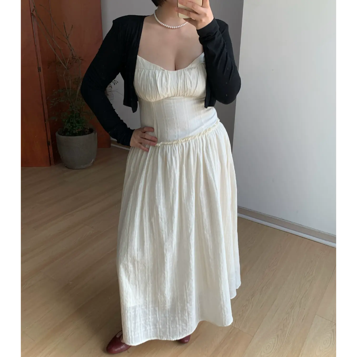 Indoor mirror styling shot of feminine textured cami maxi sundress with black cropped knit overlay, model resting one hand on hip to highlight hourglass empire waist silhouette, ruched sweetheart bust, ruffle waist seam, full flowy A-line skirt, burgundy mary jane flats, minimalist home interior